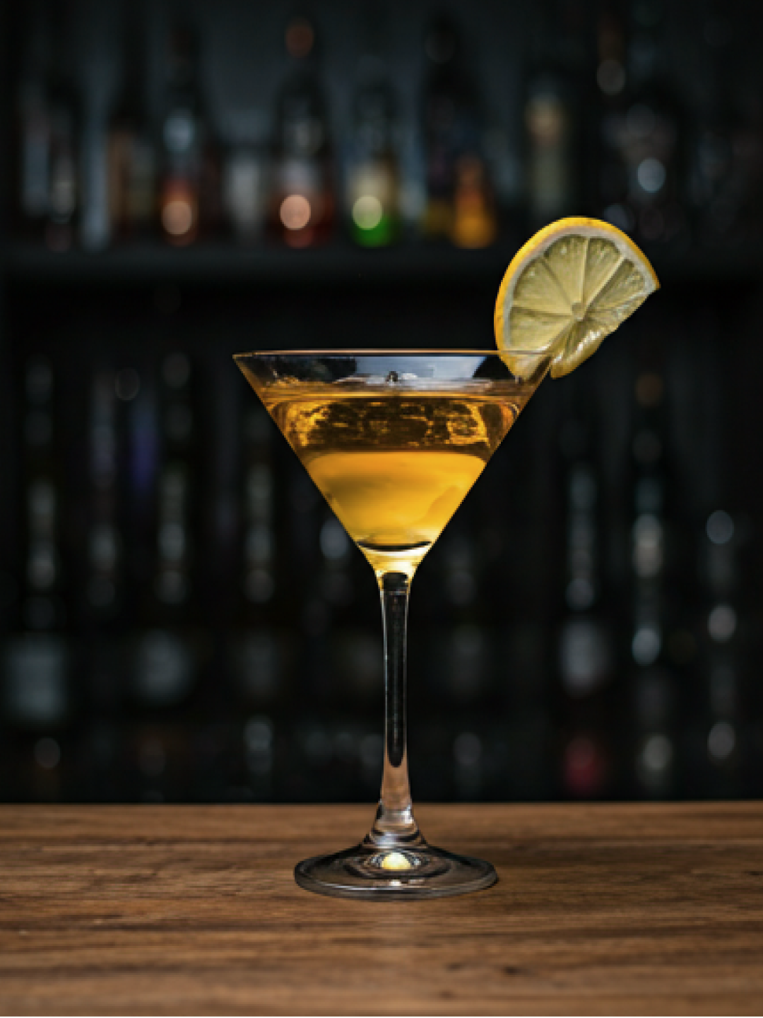A golden cocktail in a martini glass with a lemon garnish, set against a softly lit back bar.